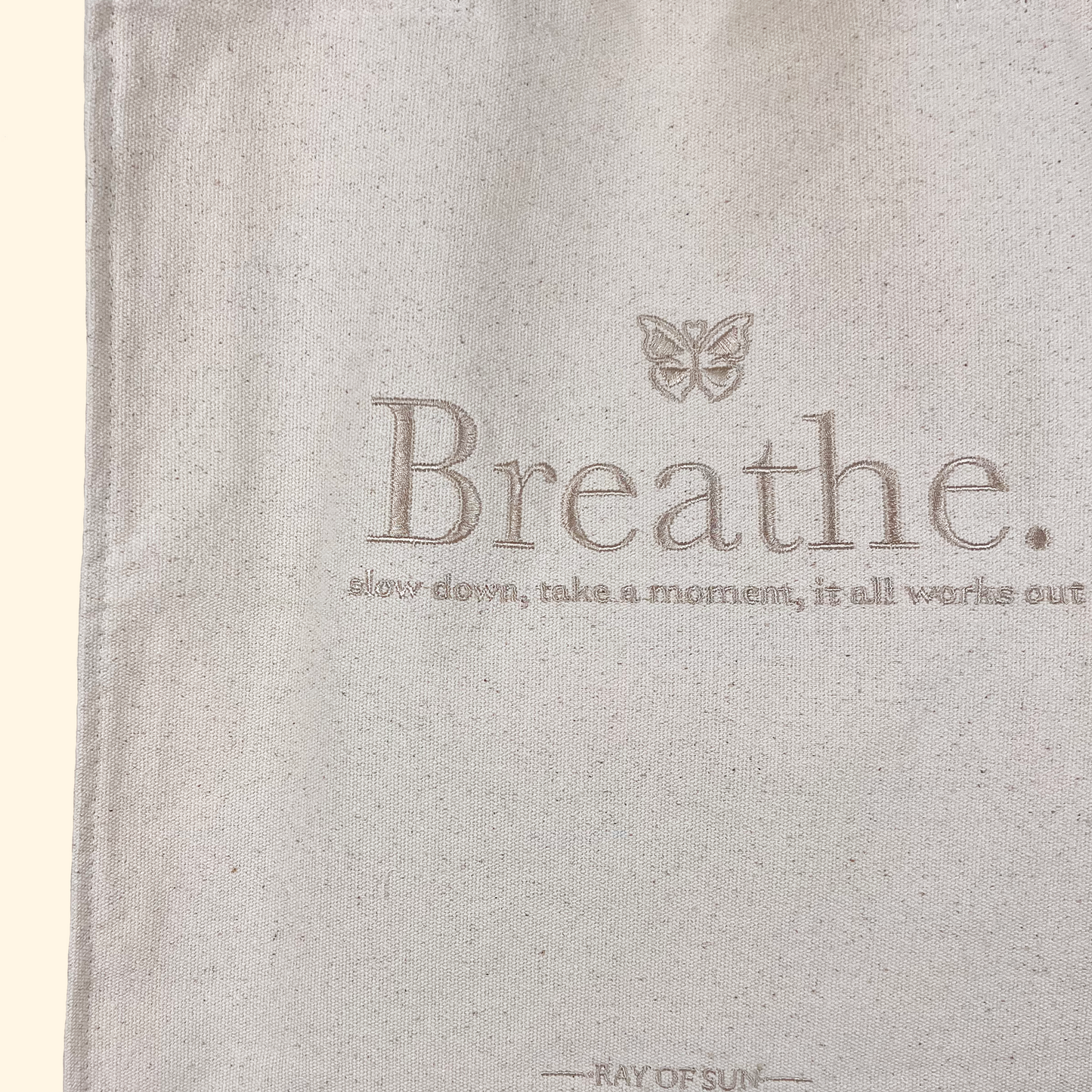 Beige tote bag closeup with silver embroidery saying "Breath" in aesthetic lettering and a butterfly on top with smaller embroidery saying " slow down, take a moment, it all works out."