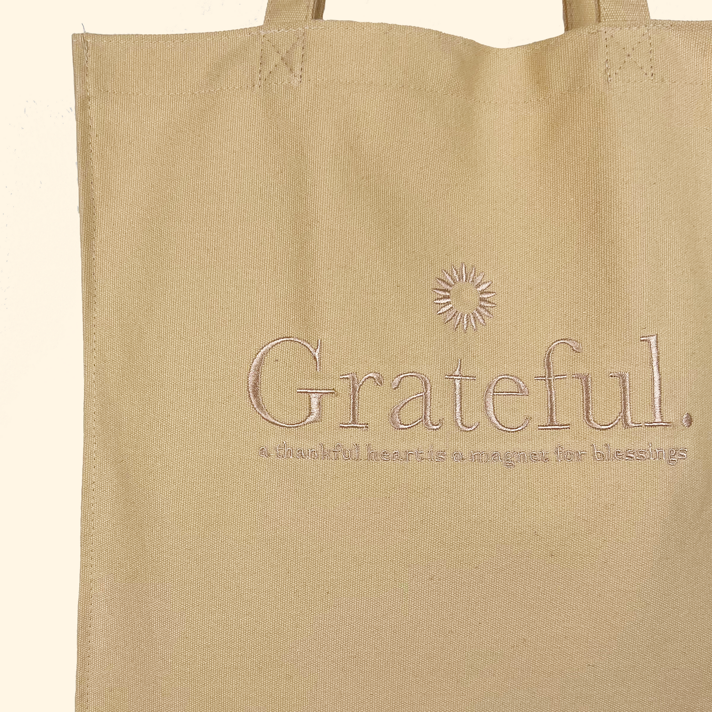 Yellow beige tote bag close up with a glistening gold embroidery saying "Grateful" and under it saying "a thankful heart is a magnet for blessing" with an embroidered sun on top 