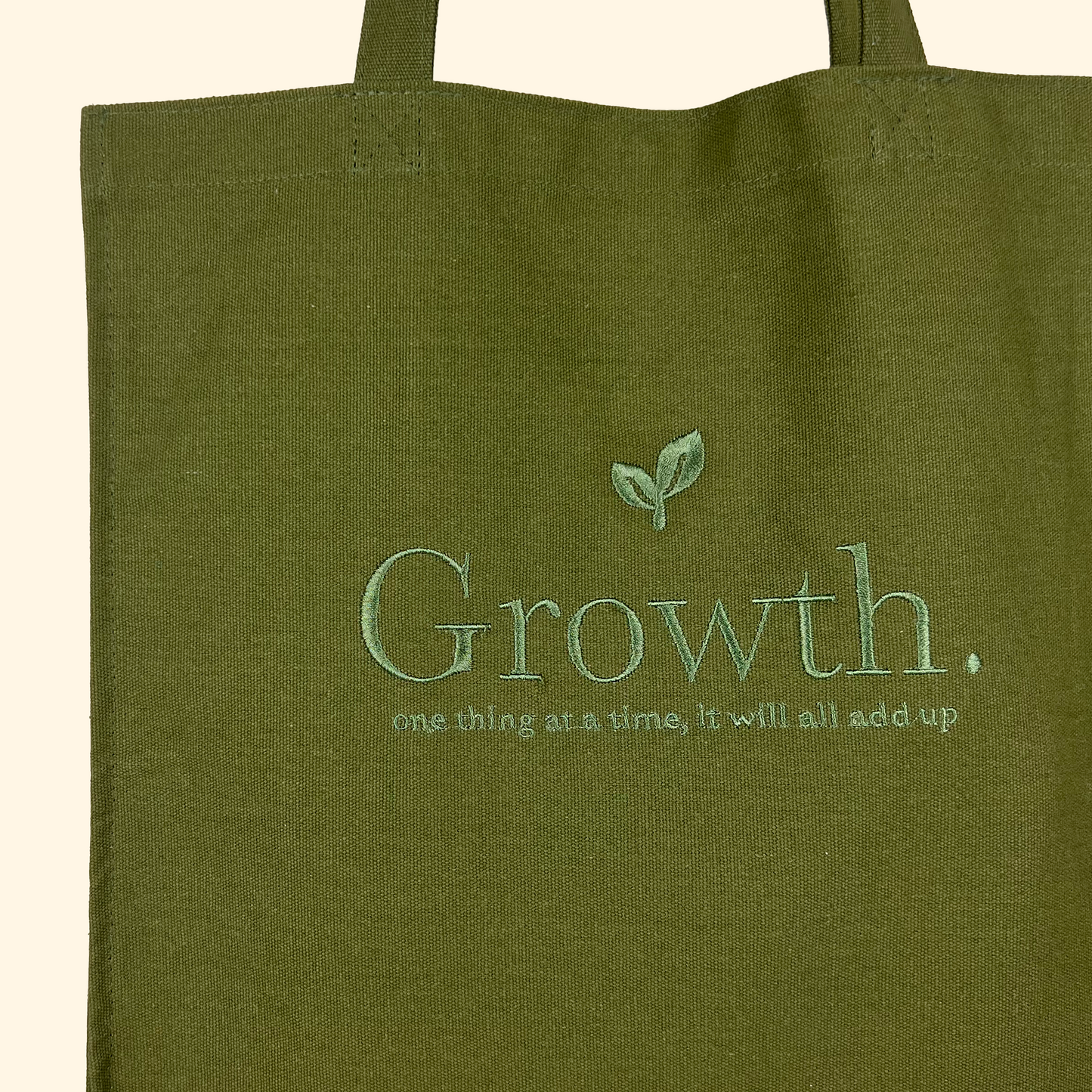 Close up of a green tote bag with embroidered letters saying "Growth" with plants embroidery and smaller embroidered text saying " one thing at a time, it will all add up."